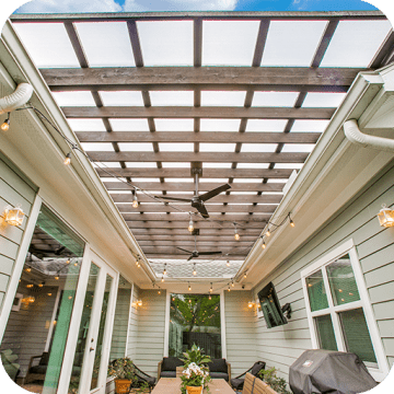 A floating pergola with ceiling fans covered with Hercules White sits over a patio within three sides of a house with windows and a french door. String lights are attached to the gutter system of the house and draped across the patio area.